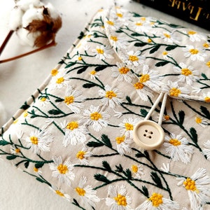 May include: A beige fabric clutch with a white daisy floral embroidery pattern. The clutch has a flap closure with a button and a white cord loop.