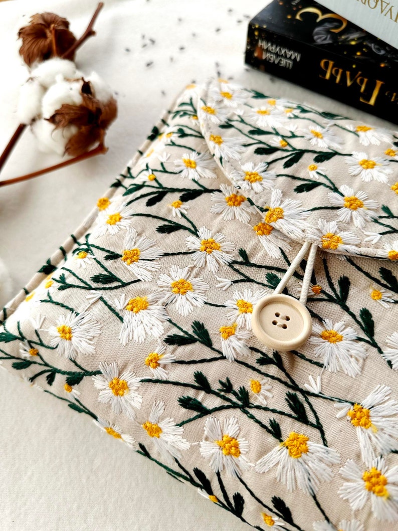May include: A beige fabric clutch with a white daisy floral embroidery pattern. The clutch has a flap closure with a button and a white cord loop.