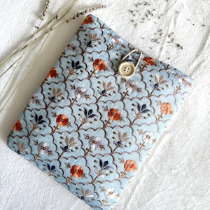 May include: A light blue case with a floral pattern and button closure. The embroidered design features orange, white, and navy blue flowers. A beige button and loop secure the case. The case is likely for a book or tablet.
