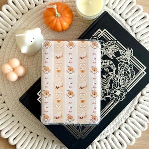 Op de afbeelding: Een doorzichtig telefoonhoesje met een herfstthema met pompoenen, boeken en strikken. De hoes ligt op een zwart boek met bloemmotieven, een witte geweven placemat en decoratieve kaarsen en pompoenen.