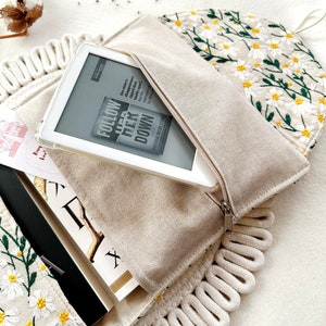 May include: A white canvas bag with a zipper closure and a white rope trim. The bag is embroidered with white daisies and green leaves. The bag is open and a white e-reader is inside. The e-reader is displaying the cover of a book titled "Follow Her Down" by Victoria Helen Stone.