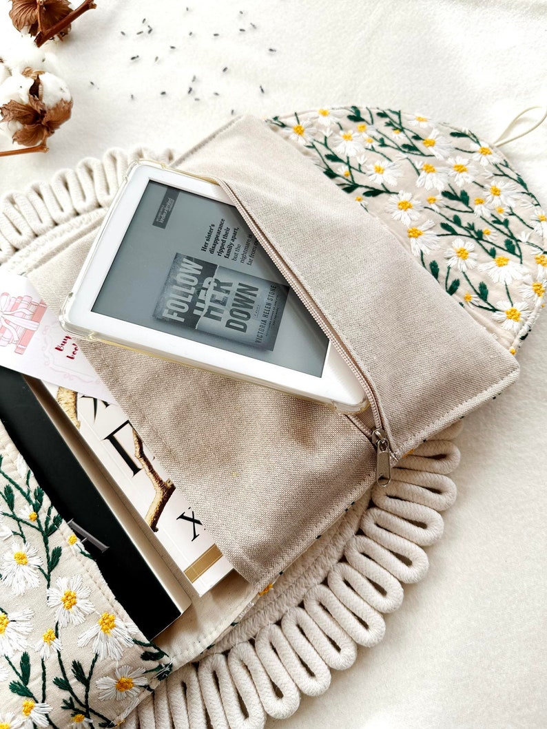 May include: A white canvas bag with a zipper closure and a white rope trim. The bag is embroidered with white daisies and green leaves. The bag is open and a white e-reader is inside. The e-reader is displaying the cover of a book titled "Follow Her Down" by Victoria Helen Stone.