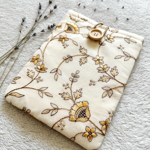 May include: A white fabric pouch with a floral embroidery design in gold and brown. The pouch has a brown button closure.