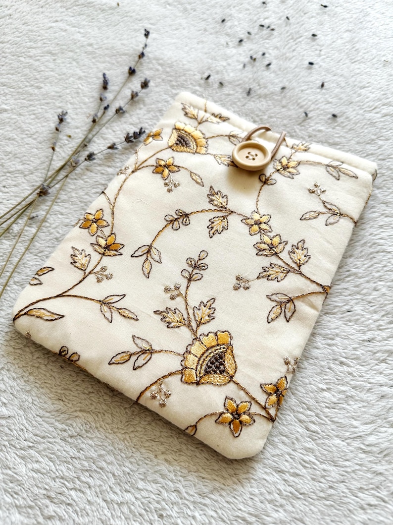 May include: A white fabric pouch with a floral embroidery design in gold and brown. The pouch has a brown button closure.