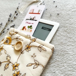 May include: A white fabric e-reader case with a floral pattern and a button closure. The case is holding a white e-reader with a bookmark that says "I have to read".