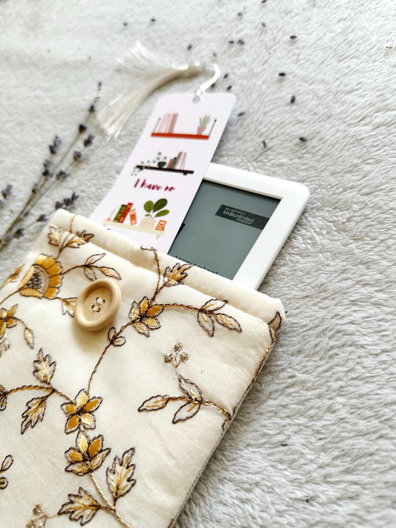 May include: A white fabric e-reader case with a floral pattern and a button closure. The case is holding a white e-reader with a bookmark that says "I have to read".