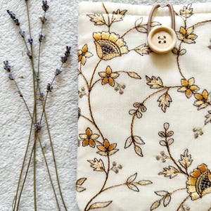 May include: A white fabric pouch with a floral pattern embroidered in gold and brown thread. The pouch has a brown button and elastic closure.