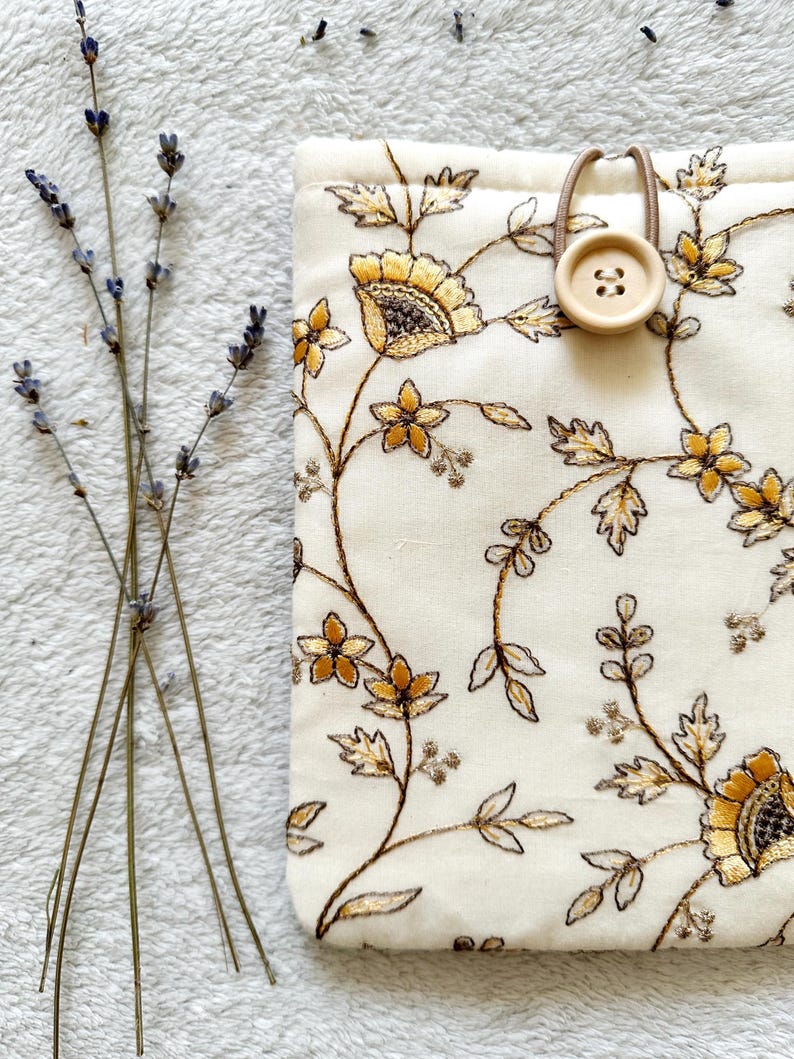 May include: A white fabric pouch with a floral pattern embroidered in gold and brown thread. The pouch has a brown button and elastic closure.