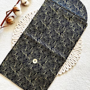 May include: A black fabric pouch with a gold cloud pattern and a single button closure. The pouch is folded in half and lying on a white surface.