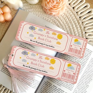 Puede incluir: Dos marcap&aacute;ginas rectangulares de papel con bordes festoneados rosas, con una nube de tormenta y un sol de dibujos animados, el texto The Grumpy x Sunshine Book Club, la frase You are the grumpy to my sunshine y un gr&aacute;fico de ticket Admit One Duo.