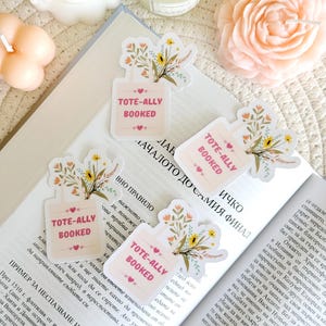 Puede incluir: Cuatro pegatinas florales con un diseño de bolsa de mano rosa y el texto "tote-ally booked" en letras blancas. Las pegatinas están en un libro con texto en un idioma extranjero.