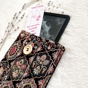 May include: A black velvet e-reader case with ornate floral embroidery in shades of pink, gold, and blue. A cream-colored button secures the flap. A guest check bookmark and an e-reader are inside the case. The bookmark reads "Buy me both flowers and books."