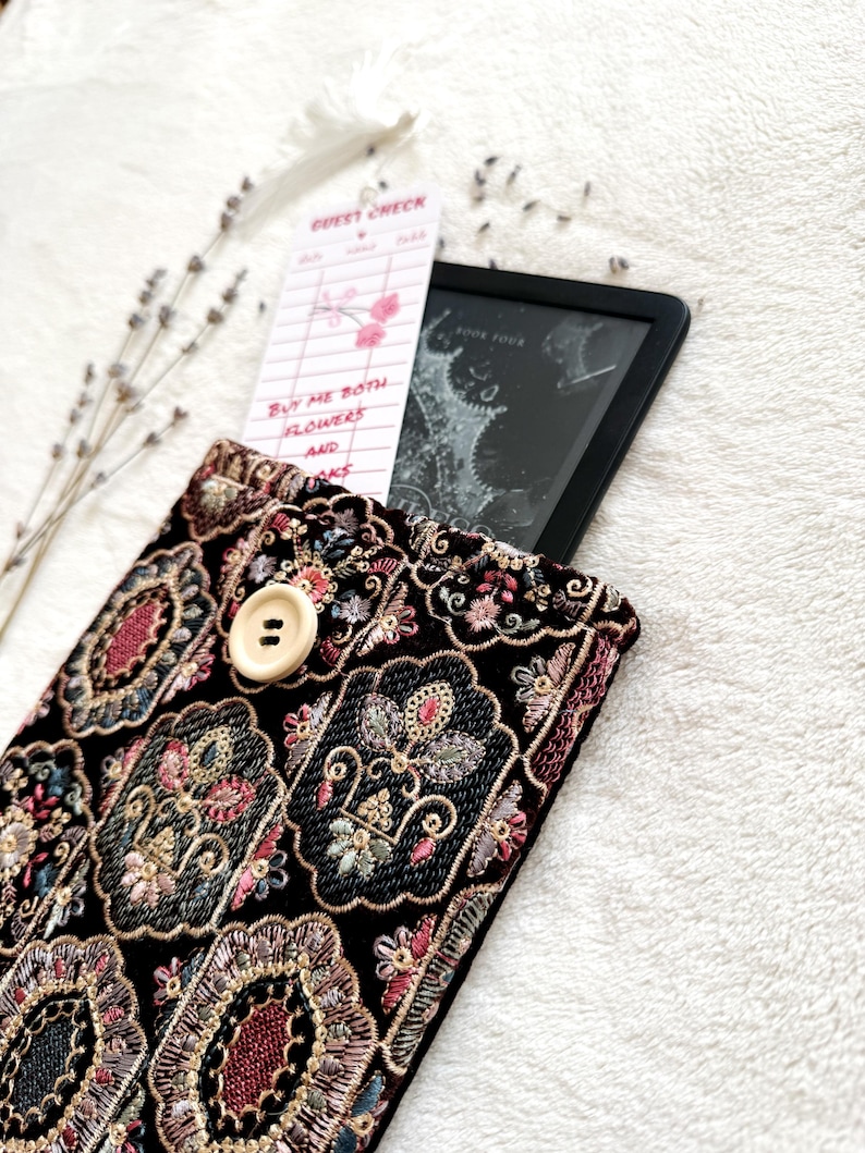 May include: A black velvet e-reader case with ornate floral embroidery in shades of pink, gold, and blue. A cream-colored button secures the flap. A guest check bookmark and an e-reader are inside the case. The bookmark reads "Buy me both flowers and books."