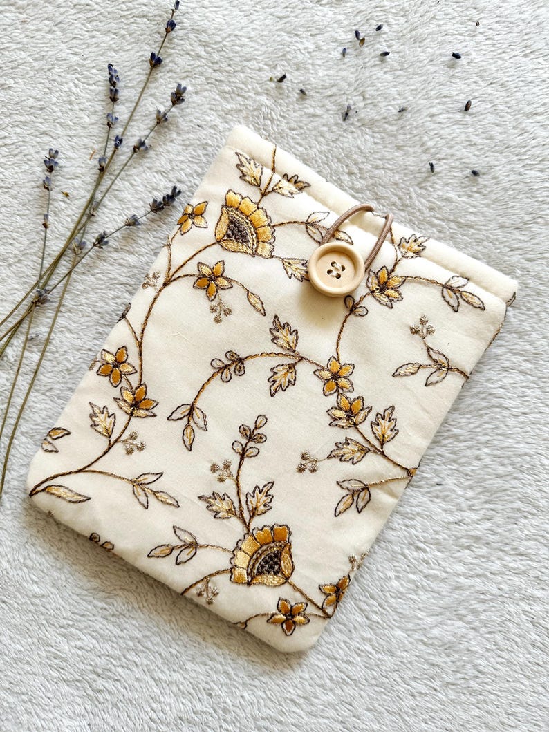 May include: A white fabric pouch with a floral embroidery design in gold thread. The pouch has a brown button closure and a brown strap.