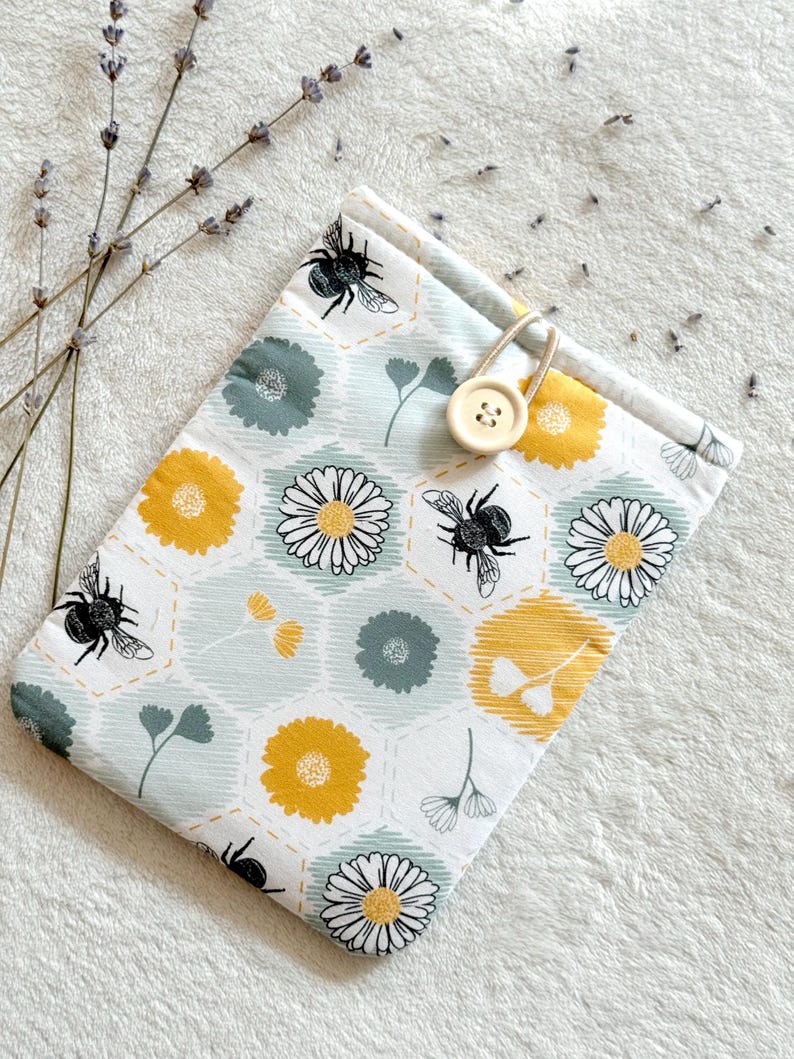 May include: A white fabric pouch with a bee and flower pattern. The pouch has a yellow button closure and a drawstring.