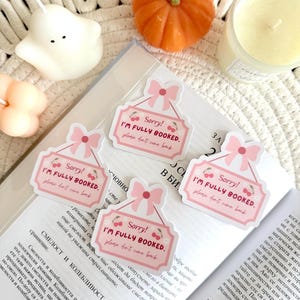 May include: Pink and white stickers with the text "Sorry! I'm Fully Booked. Please don't come back" and cherry illustrations. The stickers are on a book, with a pumpkin and candles in the background.