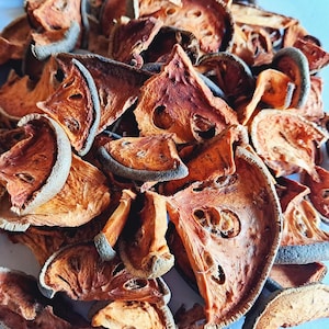 May include: A close-up shot of dried bael fruit slices. The slices are a mix of orange and brown colours, with a darker outer rind. Some slices have visible seed cavities. The texture appears dry and slightly wrinkled, suggesting dehydration.
