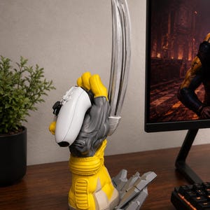 May include: A yellow and grey sculpted hand stand featuring three long silver metallic claws extending upward. The hand is gripping a white video game controller. The base of the stand is designed to look like cracked grey stone debris.