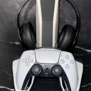 May include: A white and black gaming controller and headset stand. The stand has a white base with a black and gold accent design. A black headset rests on the stand, and a white controller is positioned at the base.