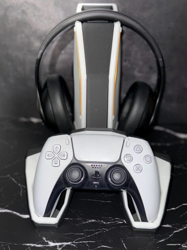 May include: A white and black gaming controller and headset stand. The stand has a white base with a black and gold accent design. A black headset rests on the stand, and a white controller is positioned at the base.