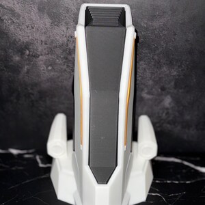 May include: A white and grey device with gold accents. The device has a rectangular shape with a black interior and two side extensions. The base is a geometric shape. The device is on a black and white surface.