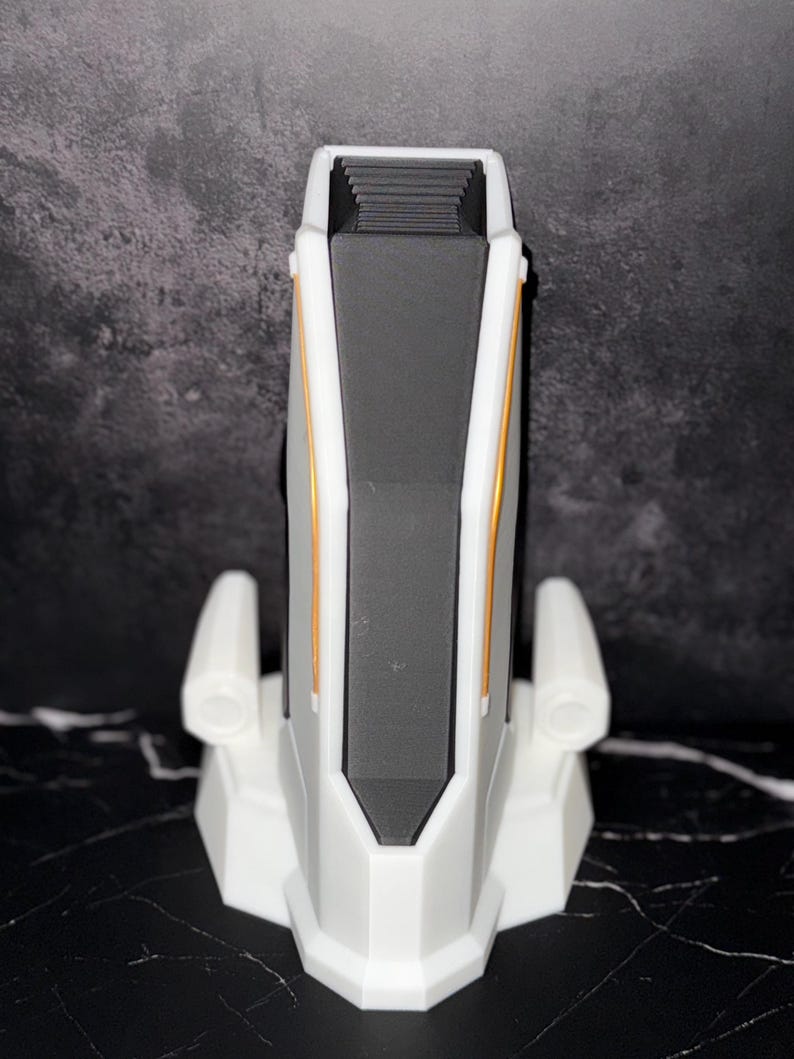 May include: A white and grey device with gold accents. The device has a rectangular shape with a black interior and two side extensions. The base is a geometric shape. The device is on a black and white surface.