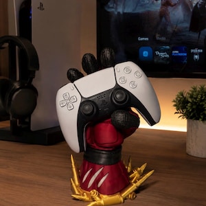 May include: A white and black wireless game controller held by a red, textured, hand-shaped stand. The base of the stand is decorated with gold-colored bullet casings. The controller features standard directional buttons, two analog sticks, and a central touchpad.