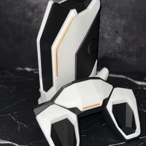 May include: A white and black device with a futuristic design. It features a rectangular base and a curved top, with orange accent lines. The geometric design suggests a tech or gaming product, with a modern aesthetic.