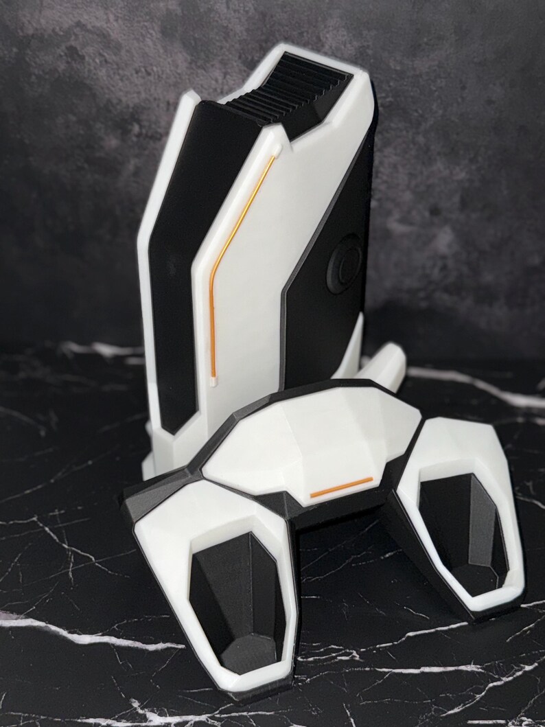 May include: A white and black device with a futuristic design. It features a rectangular base and a curved top, with orange accent lines. The geometric design suggests a tech or gaming product, with a modern aesthetic.