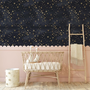 Pode incluir: Uma cena de berçário com papel de parede azul marinho com estrelas douradas. Uma moldura de parede com vieiras rosa claro fica acima de uma parede rosa claro. Um berço de vime, uma escada de madeira com um cobertor bege e uma cesta de pompons bege completam o quarto.