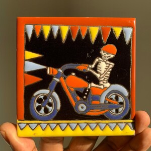 May include: A colorful ceramic tile with a black background featuring a skeleton riding a motorcycle. The motorcycle is red, orange, blue, and black. The skeleton is wearing a red helmet and has a black and white striped shirt. The tile is surrounded by a yellow, blue, and white border.