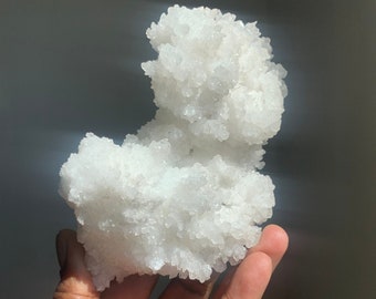 Può includere: Un grande ammasso di selenite, un tipo di gesso, di colore bianco e cristallino, con una superficie ruvida e testurizzata. L'ammasso è tenuto in una mano, mostrando la sua dimensione e la sua formazione intricata.