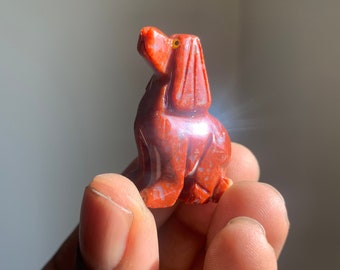 May include: A small, red, carved stone dog figurine. The dog is sitting with its head tilted up and its eyes are made of yellow stone.