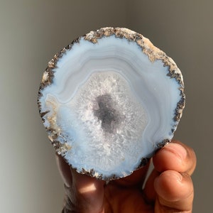 May include: A light blue agate slice with a dark grey band around the edge. The agate has a white, crystalline center.