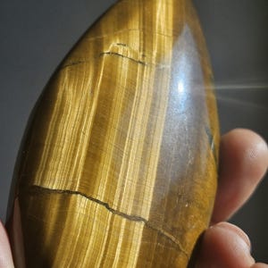 Puede incluir: Una piedra preciosa de ojo de tigre pulida, que muestra una mezcla de tonos marrón dorado y ámbar con estrías naturales. La piedra tiene una forma suave y redondeada con grietas visibles.