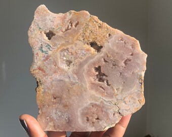 Op de afbeelding: Een grote, onregelmatig gevormde plaat van roze en bruine agaat met een natuurlijke, ruwe rand. De agaat heeft een doorschijnende, kristallijne textuur met verschillende kleine, holle holtes.