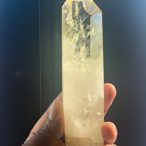 Pode incluir: Um grande cristal de quartzo transparente com uma ponta. O cristal tem um tom amarelo p&aacute;lido e &eacute; mantido contra um fundo escuro. A superf&iacute;cie do cristal tem fraturas e inclus&otilde;es internas. Um mineral natural.
