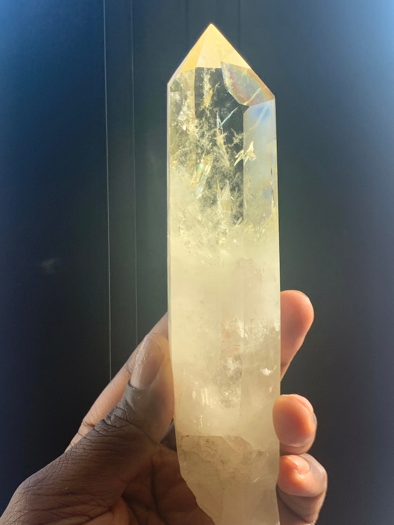 Pode incluir: Um grande cristal de quartzo transparente com uma ponta. O cristal tem um tom amarelo p&aacute;lido e &eacute; mantido contra um fundo escuro. A superf&iacute;cie do cristal tem fraturas e inclus&otilde;es internas. Um mineral natural.