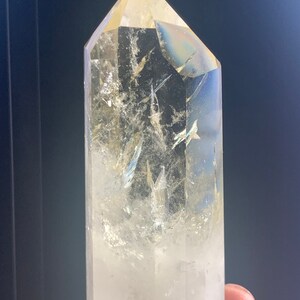 Pode incluir: Uma ponta de cristal de quartzo transparente com uma parte superior facetada. O cristal tem fraturas e inclus&otilde;es internas e um ligeiro tom amarelo. O cristal &eacute; mantido contra um fundo escuro.