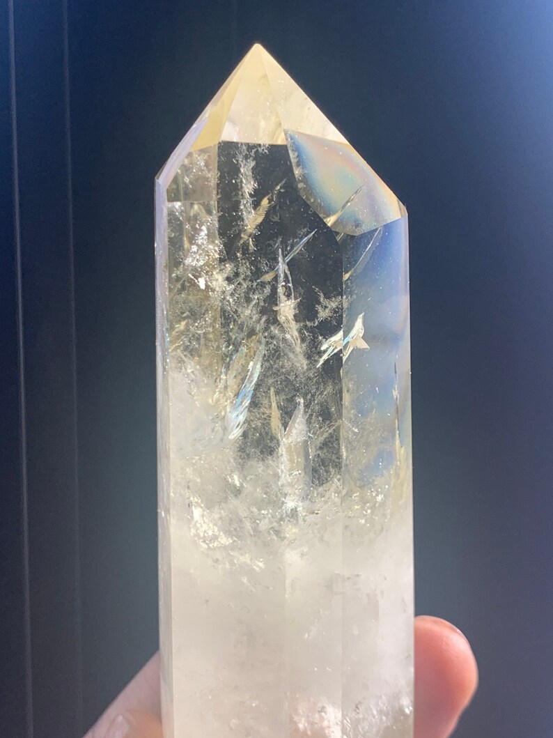 Pode incluir: Uma ponta de cristal de quartzo transparente com uma parte superior facetada. O cristal tem fraturas e inclus&otilde;es internas e um ligeiro tom amarelo. O cristal &eacute; mantido contra um fundo escuro.