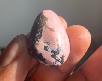 May include: A pink and black polished gemstone with a smooth, rounded shape. The stone has a unique pattern of pink and black swirls.