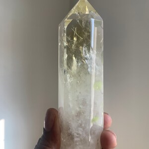 Pode incluir: Uma ponta de cristal de quartzo transparente com um tom amarelo no topo. O cristal &eacute; segurado em uma m&atilde;o e possui inclus&otilde;es internas. O cristal &eacute; uma pedra preciosa natural e polida.