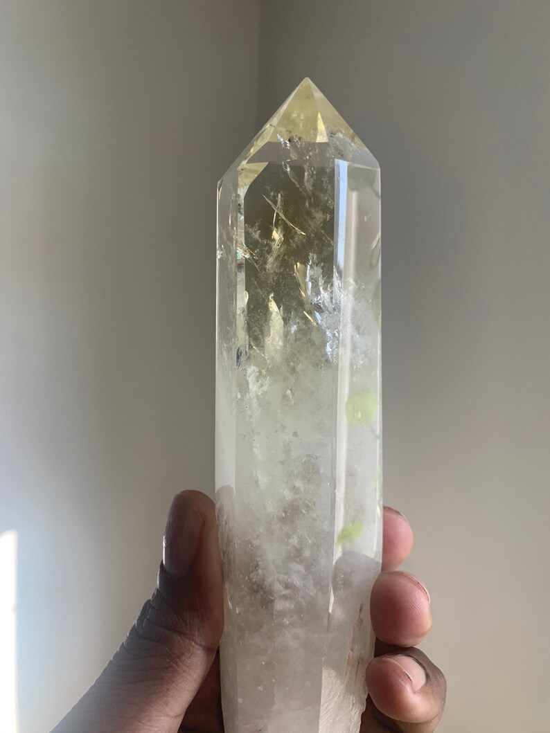 Pode incluir: Uma ponta de cristal de quartzo transparente com um tom amarelo no topo. O cristal &eacute; segurado em uma m&atilde;o e possui inclus&otilde;es internas. O cristal &eacute; uma pedra preciosa natural e polida.
