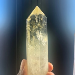 Pode incluir: Uma ponta de cristal de quartzo amarelo claro com um design facetado. O cristal &eacute; segurado em uma m&atilde;o e tem um topo pontiagudo. O cristal tem uma apar&ecirc;ncia nublada e texturizada e &eacute; de cor amarelo claro.