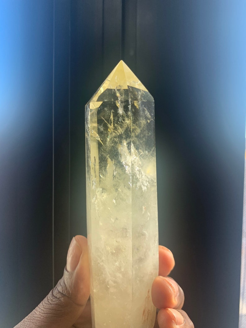 Pode incluir: Uma ponta de cristal de quartzo amarelo claro com um design facetado. O cristal &eacute; segurado em uma m&atilde;o e tem um topo pontiagudo. O cristal tem uma apar&ecirc;ncia nublada e texturizada e &eacute; de cor amarelo claro.