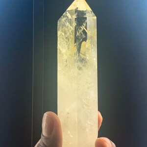 Pode incluir: Uma ponta de cristal de citrino amarelo claro. O cristal &eacute; segurado em uma m&atilde;o e tem um topo facetado e pontiagudo. O cristal &eacute; de cor amarelo p&aacute;lido e tem inclus&otilde;es internas. O fundo &eacute; de cor escura e s&oacute;lida.