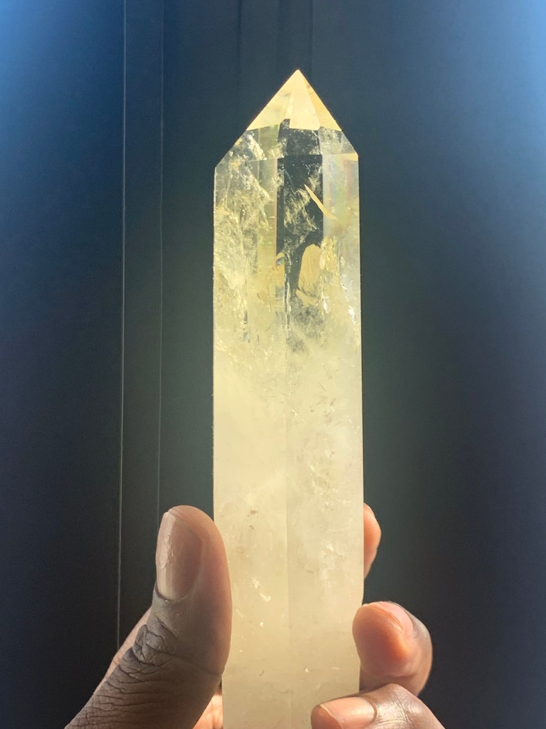 Pode incluir: Uma ponta de cristal de citrino amarelo claro. O cristal &eacute; segurado em uma m&atilde;o e tem um topo facetado e pontiagudo. O cristal &eacute; de cor amarelo p&aacute;lido e tem inclus&otilde;es internas. O fundo &eacute; de cor escura e s&oacute;lida.
