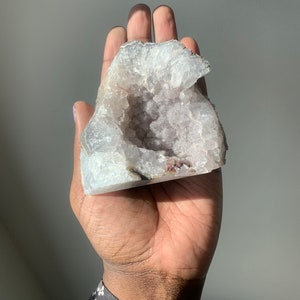 K&ouml;nnte beinhalten: Ein gro&szlig;er, wei&szlig;er Geodenkristall mit einer hohlen Mitte, die mit kleineren, violetten Kristallen gef&uuml;llt ist. Die Geode wird in einer Hand gehalten.