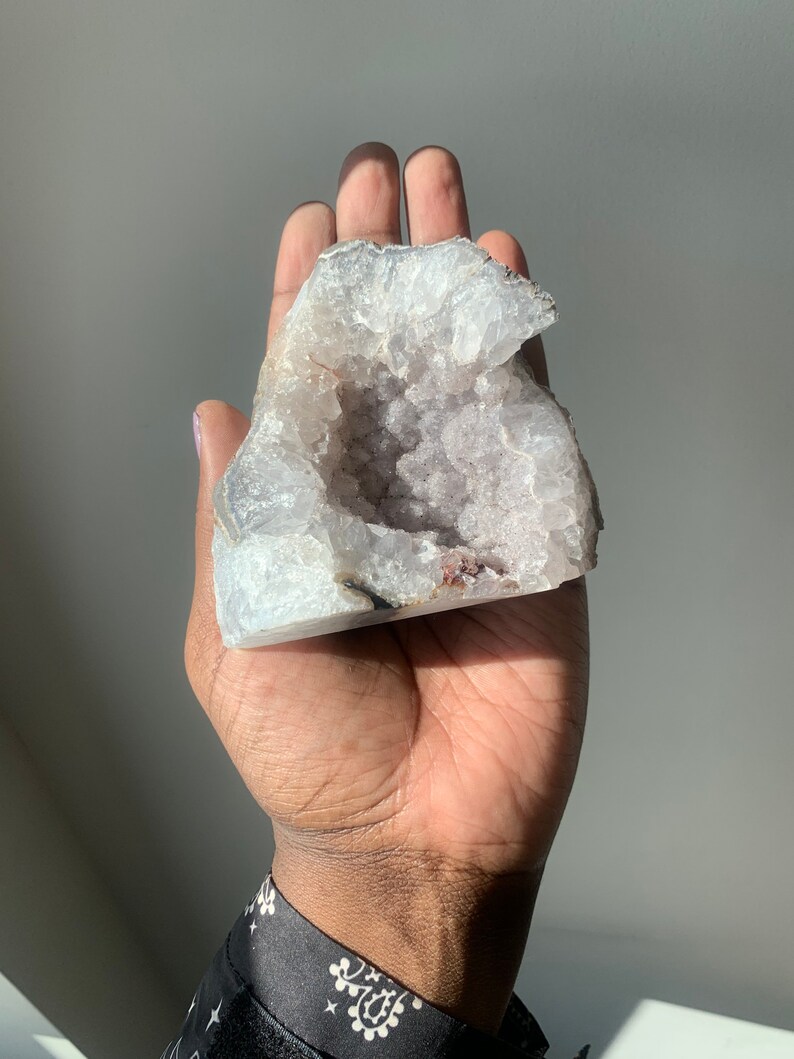 K&ouml;nnte beinhalten: Ein gro&szlig;er, wei&szlig;er Geodenkristall mit einer hohlen Mitte, die mit kleineren, violetten Kristallen gef&uuml;llt ist. Die Geode wird in einer Hand gehalten.