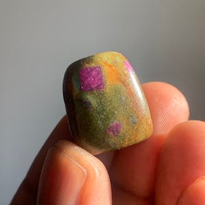 May include: A tumbled gemstone with a mix of colors, including green, orange, and pink. The stone has a smooth, rounded shape and appears to be a natural mineral specimen. The pink areas are likely ruby inclusions.